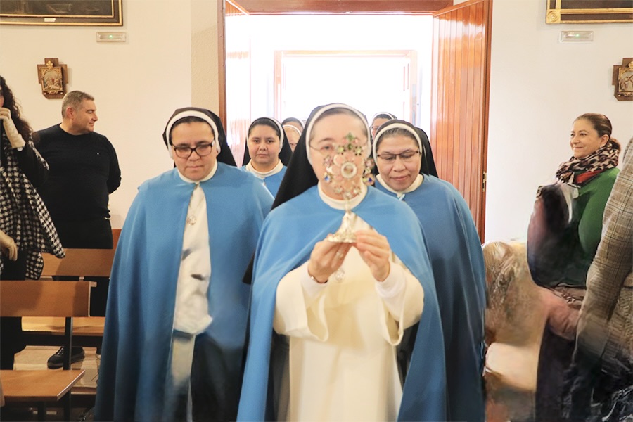 Una monja con su hábito azul y blanco camina en procesión con una reliquia de plata en sus manos. Al fondo otras monjas le acompañan.